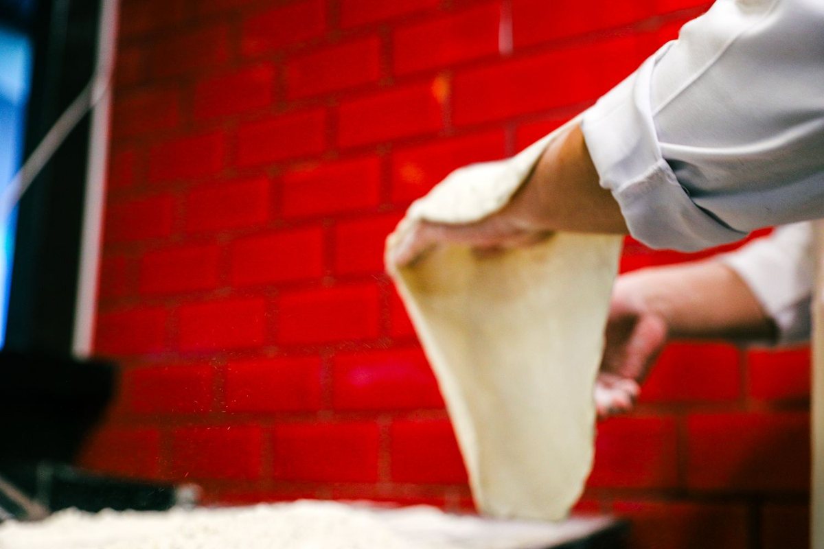 A cook stretching pizza dough by hand against a red brick wall