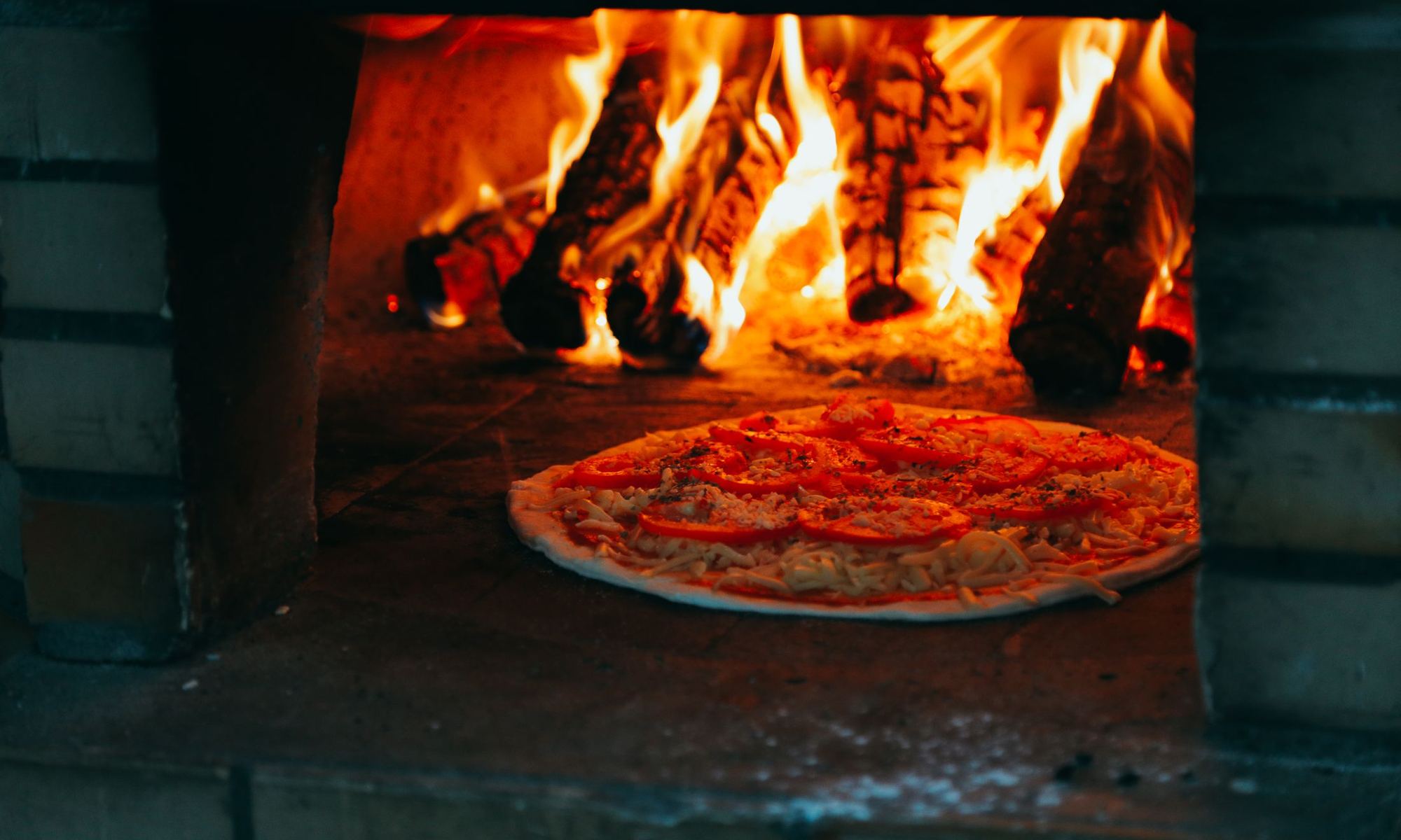 A pizza on the brick floor of a wood-fired oven with logs burning behind it