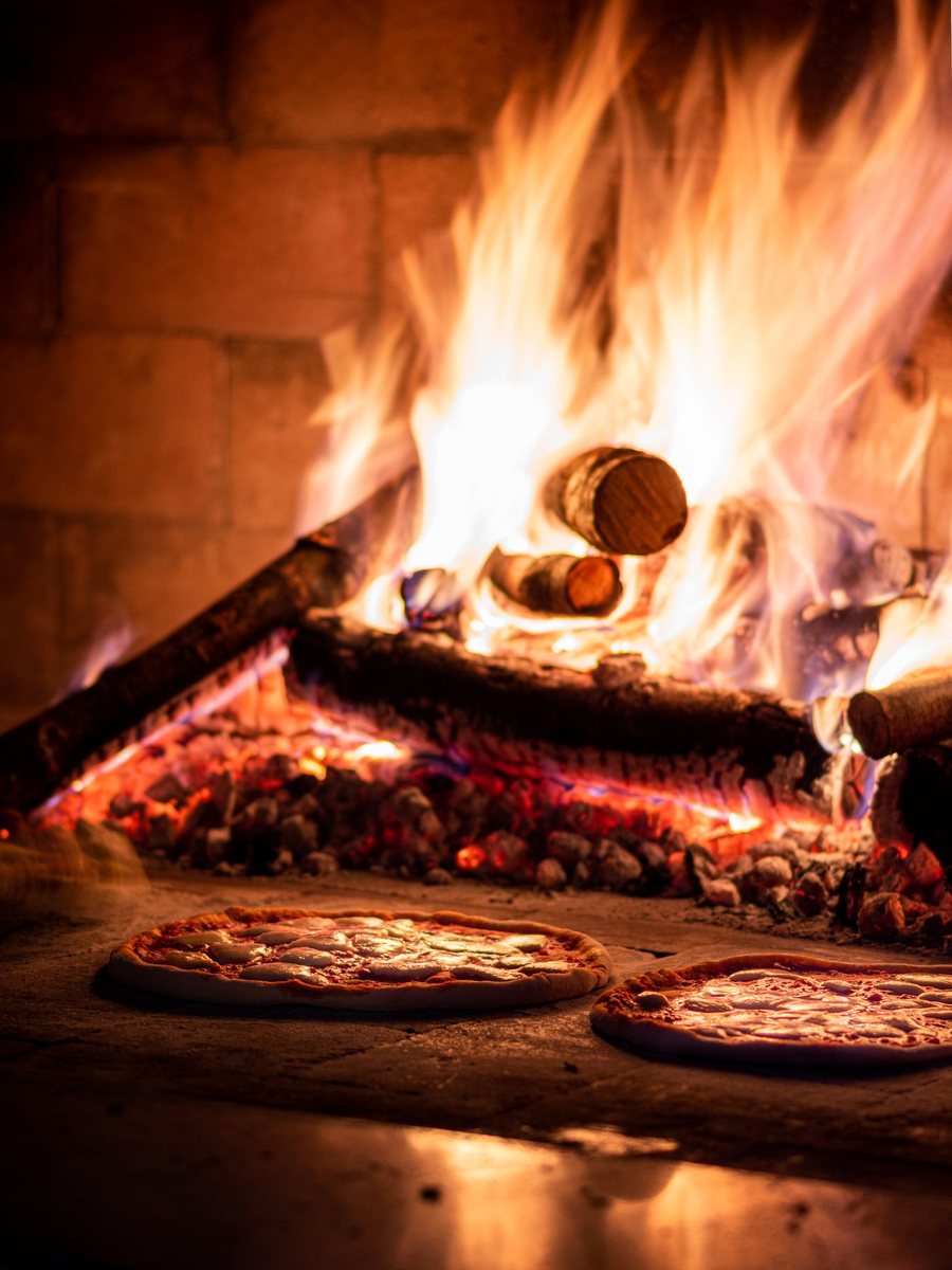 Logs burning at the back of a brick pizza oven above a bed of glowing coals