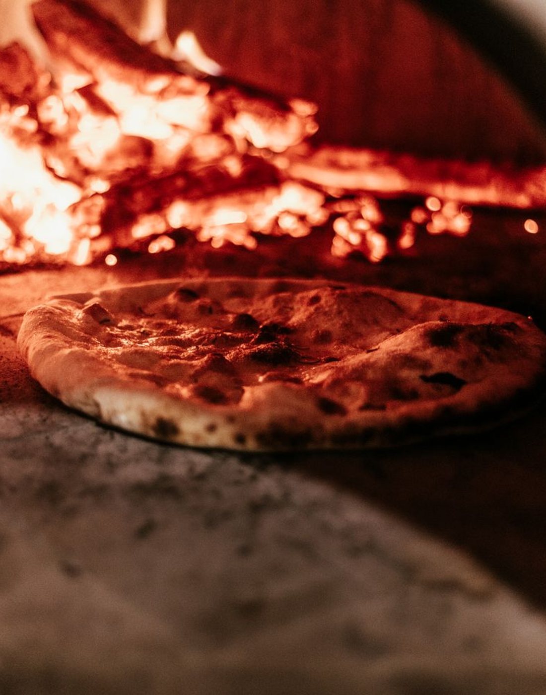 A pizza baking on the stone floor of a wood-fired oven beside burning logs